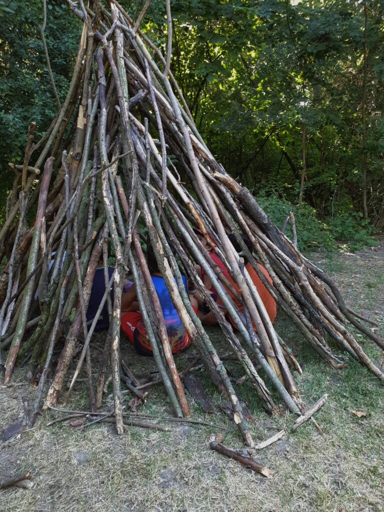 bambini e bambine che giocano all'aperto dentro a una tenda costruita con tronchi di alberi
